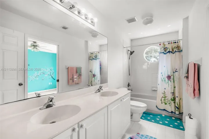 Double vanity with integrated white countertops and dual under-mount sinks, expansive wall-to-wall vanity mirror, overhead vanity lighting, circular accent window, and white tile tub surround