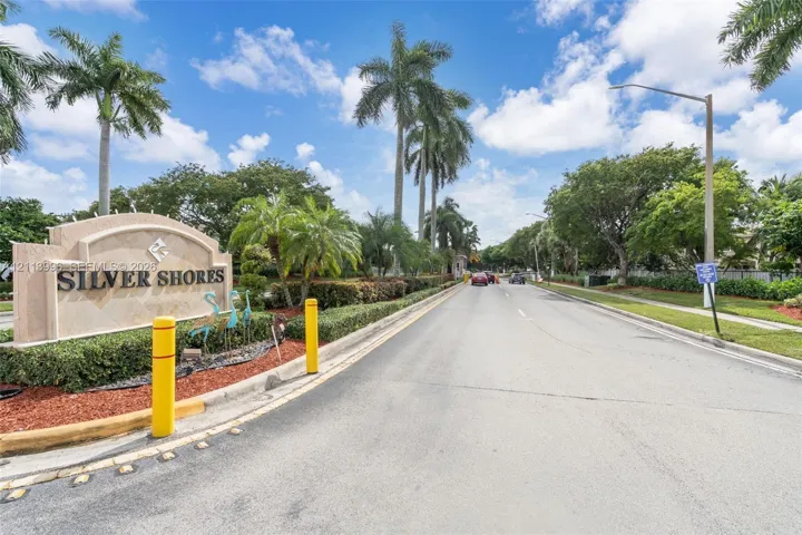 Community entrance monument featuring stone-finish masonry, mature palm trees, landscaped shrubbery, and paved roadway access