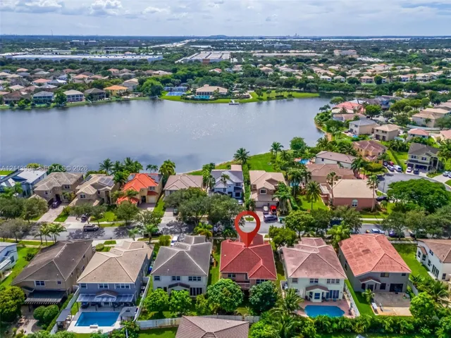 Aerial perspective of a multi-story residence with a multi-gabled tile roof, situated in a suburban community featuring a large lake and mature landscaping