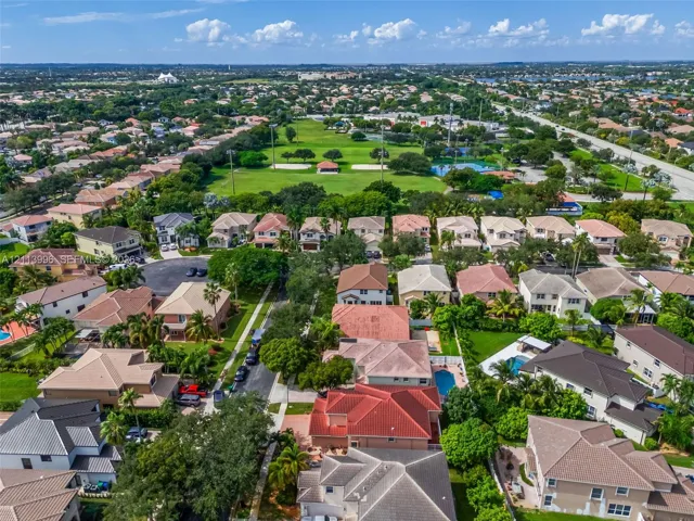 Aerial perspective of a suburban neighborhood featuring paved streets, mature landscaping, multi-story residences with tiled roofs, and an expansive recreational green space