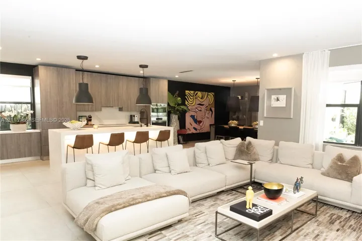 Open-concept interior featuring light wood-finish cabinetry, a large kitchen island with integrated under-counter lighting, stone slab surfacing, and matte black pendant lighting