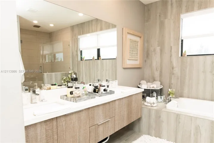 Floating wood-finish vanity with dual integrated sinks and polished chrome fixtures, oversized wall-to-wall mirror, built-in soaking tub, and tile-wrapped walls