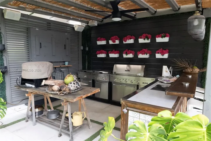 Covered outdoor kitchen featuring a multi-burner stainless steel grill, sleek cabinetry, integrated sink, and dark wood-finish paneled wall with decorative wall-mounted planters
