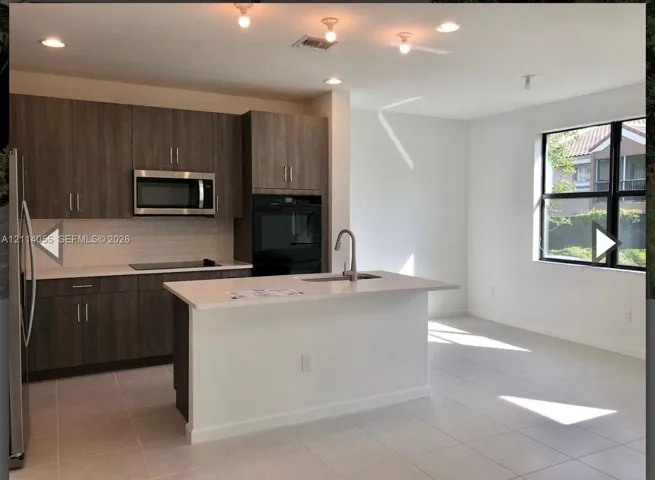 Kitchen featuring wood-finish cabinetry, a center island with a stone-surface countertop, integrated stainless steel wall oven and microwave, subway tile backsplash, and ceramic tile flooring