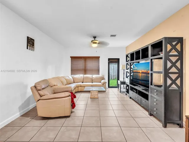 Spacious living area featuring large-format ceramic tile flooring, built-in floor-to-ceiling wooden shelving unit, ceiling fan with integrated lighting, and horizontal window blinds