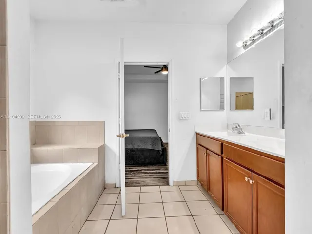 Bathroom featuring a built-in soaking tub with neutral tile surround, a wood-finish dual vanity with a white countertop, and a full-width wall-mounted mirror