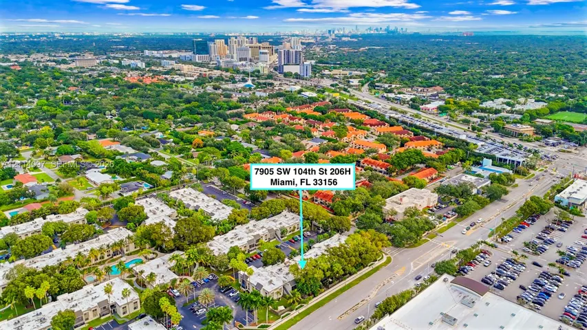 Aerial perspective of residential community featuring low-rise multi-family structures, mature tree canopy, integrated parking areas, and proximity to regional infrastructure and urban skyline