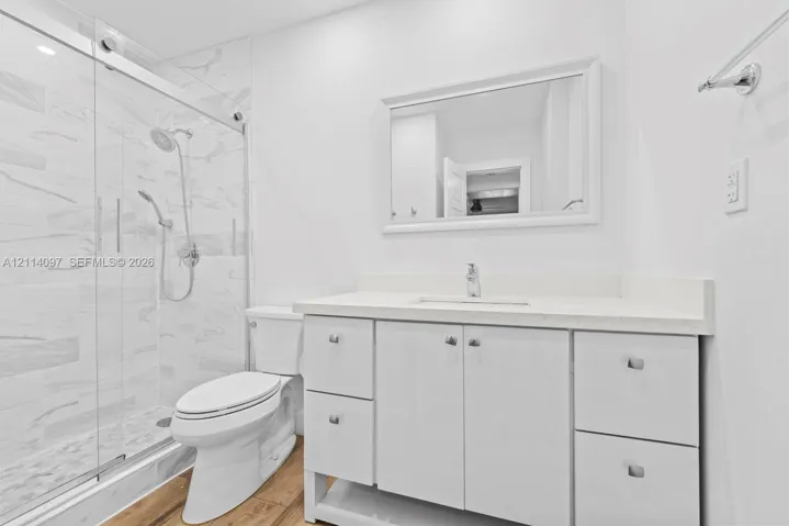 Bathroom featuring a white vanity with integrated basin and stone-finish countertop, a glass-enclosed walk-in shower with tiled walls, and wood-finish flooring