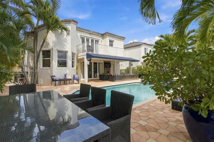 Stucco exterior featuring a second-story balcony with metal railings, a paved stone patio, an in-ground swimming pool, and a retractable awning