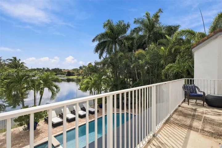 Second-story balcony with white metal railings overlooking a rectangular swimming pool, paver patio, mature palm trees, and a pond