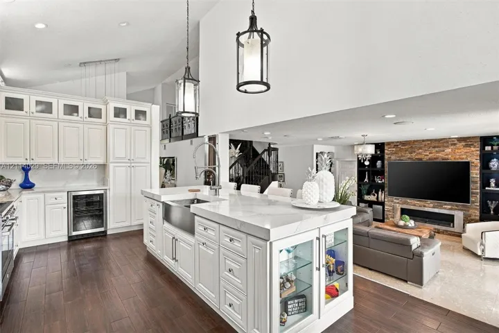 Kitchen featuring white shaker-style cabinetry, an expansive center island with an integrated farmhouse sink, glass-front display shelving, stone-veneer fireplace surround, and dark wood-finish flooring