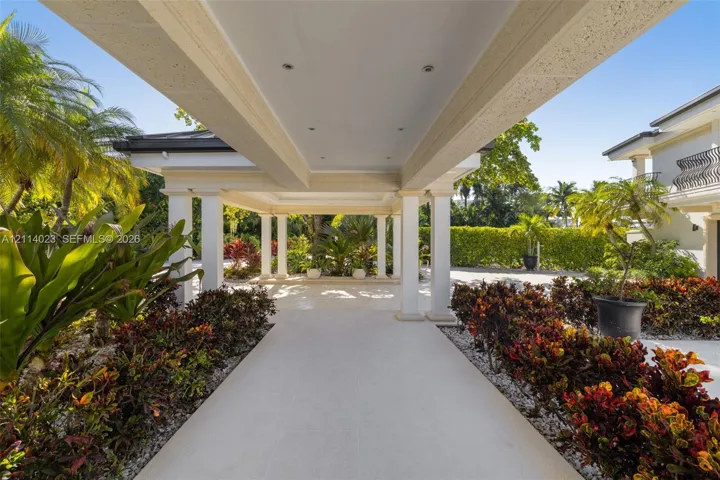 Covered porte-cochère featuring a light-toned stone tile surface, structural white columns, recessed lighting, and surrounding professional landscaping