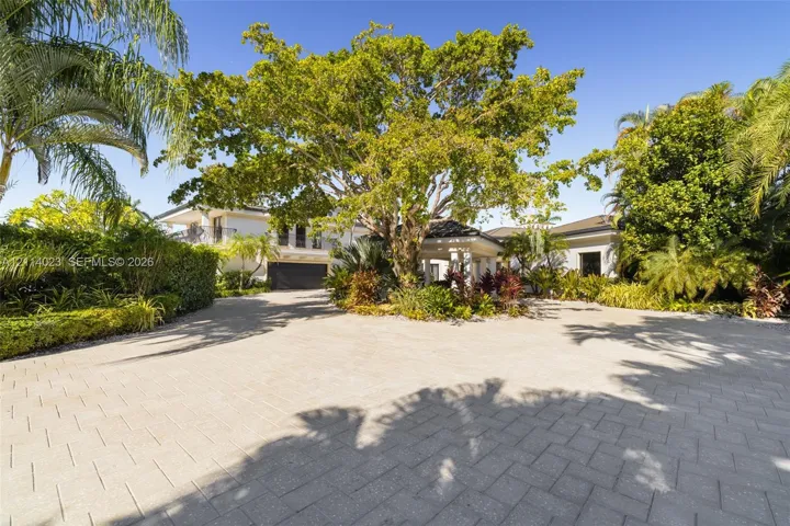 Paver driveway leading to a covered entryway, mature specimen trees, and established perimeter landscaping