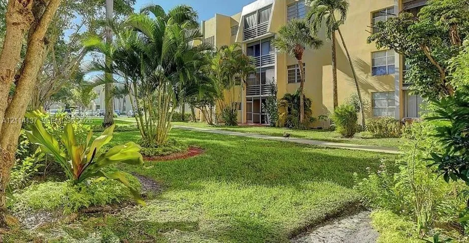Multi-story building exterior featuring smooth stucco siding, aluminum-frame windows, and screened balconies, set amidst landscaped grounds with mature palms and established greenery