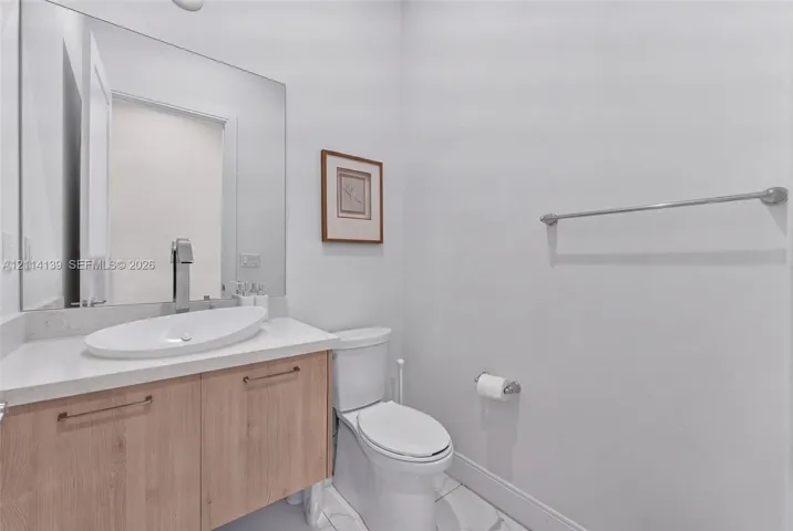 Wood-finish vanity cabinet, white oval vessel sink, light-toned countertop, large frameless mirror, and dual-flush toilet