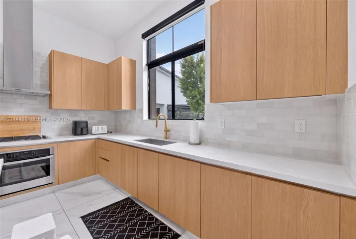 Kitchen featuring wood-finish flat-panel cabinetry, light-toned stone-surface countertops, white subway tile backsplash, stainless steel range with integrated vent hood, and brass-finish faucet hardware