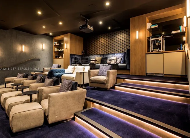 Media room featuring tiered flooring with integrated LED lighting, built-in wooden cabinetry, custom textured wall paneling, and an overhead projector mount