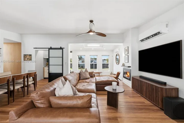 Open-concept interior featuring wood-finish flooring, a contemporary ceiling fan, a wall-mounted fireplace, a black-framed sliding barn door with frosted panels, and a wall-mounted mini-split HVAC unit