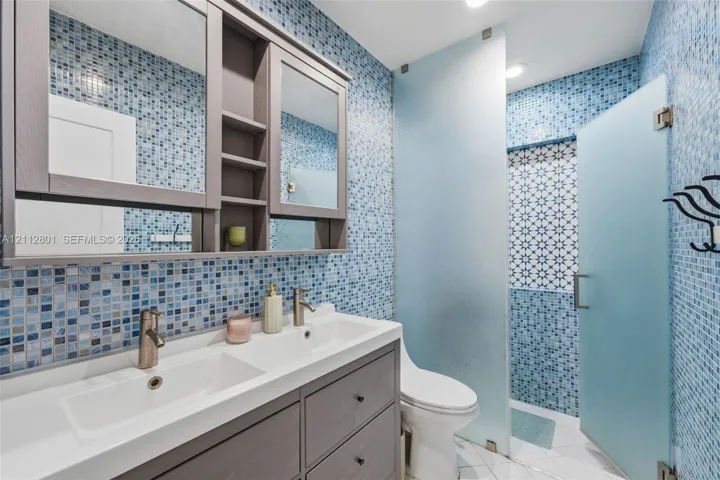 Double vanity featuring a white integrated countertop with dual basins and matte-finish hardware, built-in medicine cabinets, full-height blue glass mosaic tile walls, and a walk-in shower enclosure with frosted glass panels