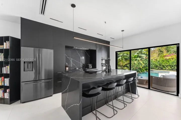 Contemporary kitchen featuring matte black cabinetry, waterfall island with stone-finish surfaces, integrated stainless steel appliances, and large-format sliding glass doors