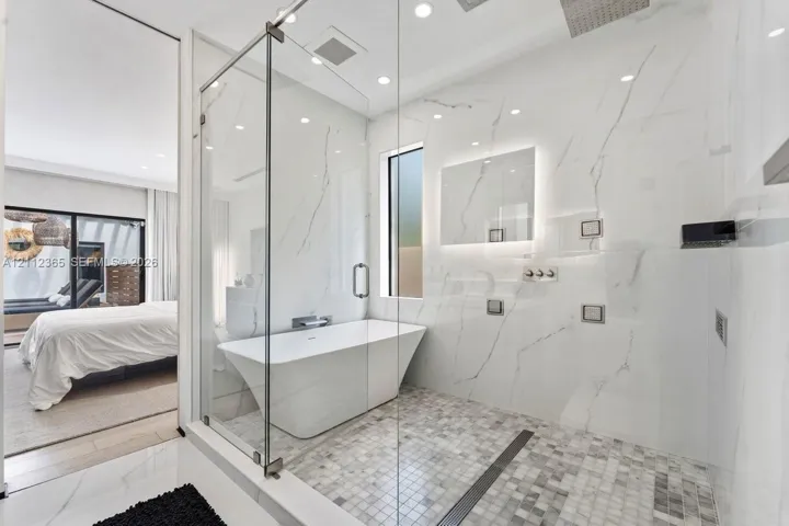 Walk-in wet room featuring a freestanding soaking tub, frameless glass enclosure, floor-to-ceiling stone-finish wall tile, mosaic tile flooring, and wall-mounted plumbing fixtures