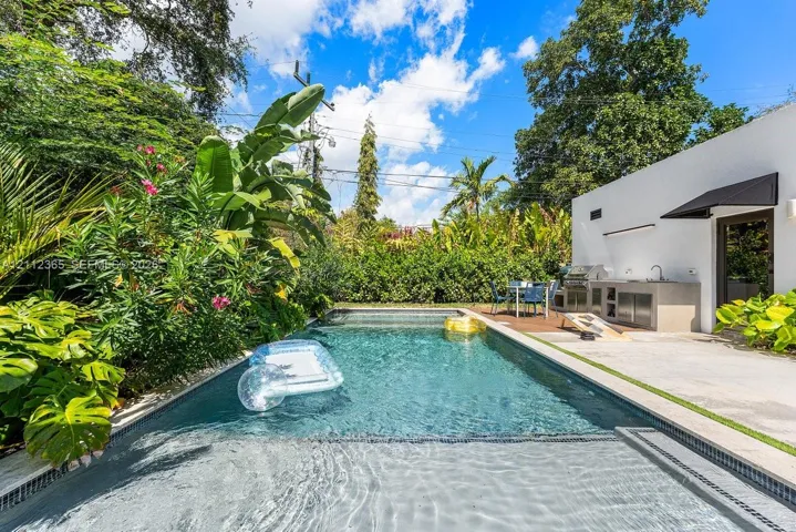 Swimming pool with a baja shelf, concrete patio, built-in outdoor kitchen with sink, and lush perimeter landscaping