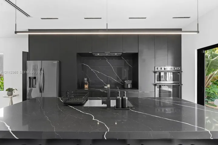 Kitchen featuring matte dark cabinetry, expansive dark stone-patterned island with matching backsplash, linear pendant lighting, and stainless steel built-in appliances