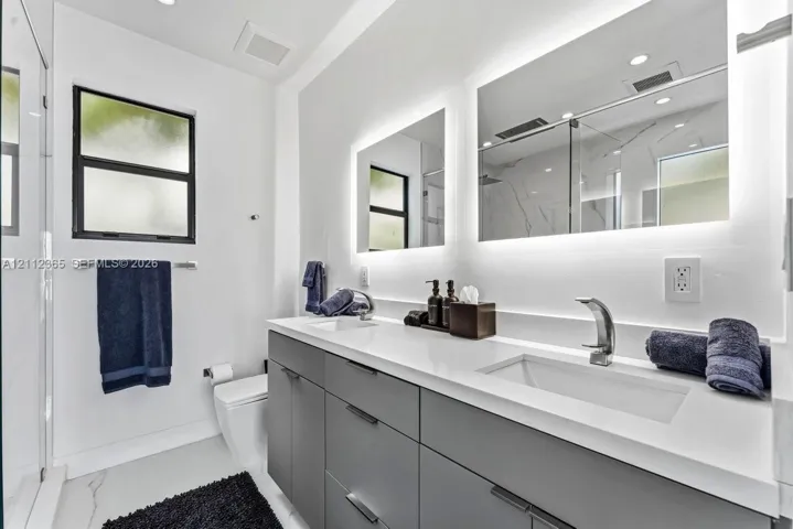 Double vanity with grey cabinetry, rectangular undermount sinks, quartz countertops, and back-lit mirrors, complemented by a glass-enclosed shower featuring large-format tile surrounds