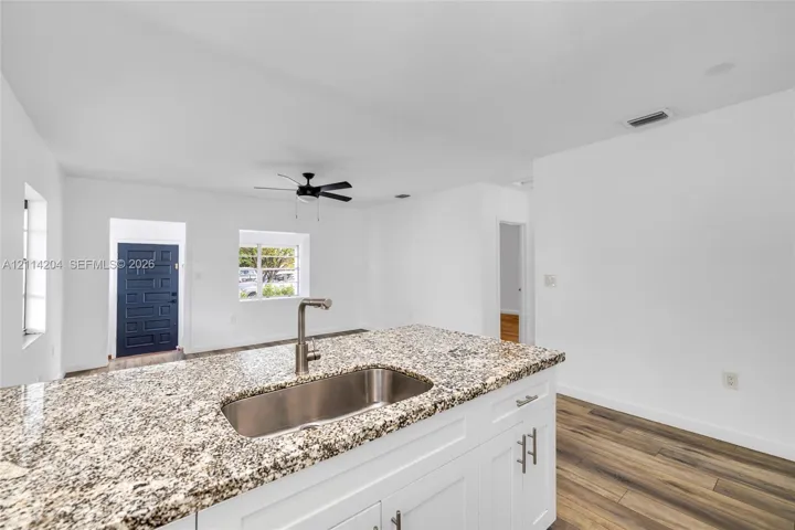 Kitchen island featuring stone-surfaced countertops, stainless steel undermount sink, gooseneck faucet, white shaker-style cabinetry, and wood-finish flooring