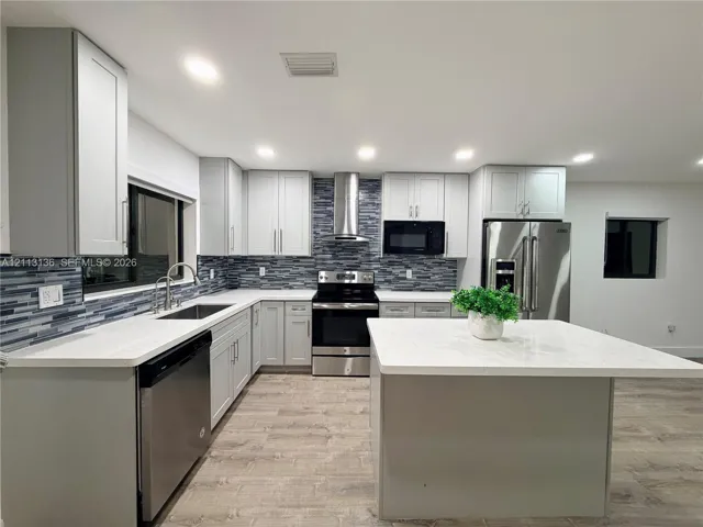 Kitchen featuring shaker-style cabinetry, a central island with a solid surface countertop, stainless steel appliances, a multi-tonal tile backsplash, and wood-finish flooring