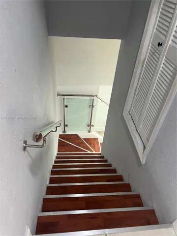 Staircase featuring wood-finish treads, metal nosing, wall-mounted handrail, louvered closet doors, and glass panel railing with metal posts