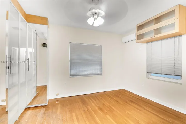 Room featuring wood-finish flooring, built-in floor-to-ceiling wardrobe cabinetry with mirrored doors, wall-mounted ductless climate control unit, and overhead ceiling fan with light fixture