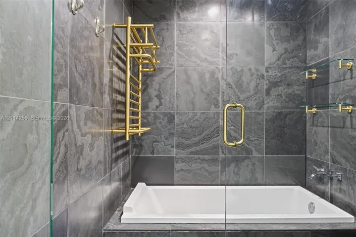 Large-format charcoal stone tile surround with a white deep-soaking tub, brass-finish heated towel rack, wall-mounted faucet, and glass shelving