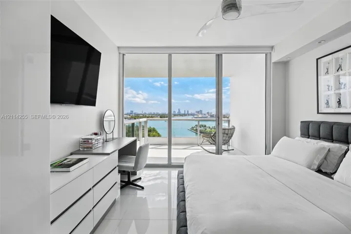Floor-to-ceiling sliding glass doors, white tile flooring, recessed ceiling lighting, three-blade ceiling fan, and private balcony with water and city skyline backdrop