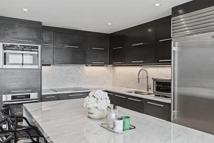 Custom dark-finish cabinetry featuring horizontal bar pulls, expansive stone-surface center island, integrated stainless steel refrigeration, built-in wall oven and microwave combination, and stone-surface backsplash with under-cabinet lighting