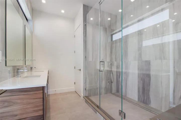 Dual-sink vanity with wood-finish cabinetry and white countertops, walk-in shower with floor-to-ceiling tile, frameless glass enclosure, and recessed lighting