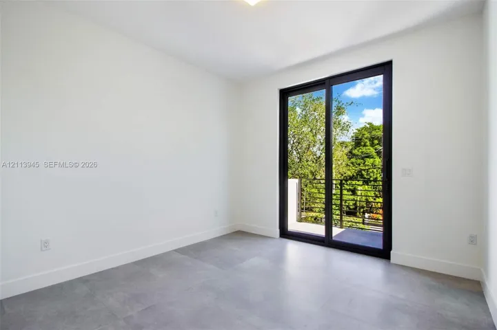 Interior room featuring large-format grey tile flooring, minimalist white walls, and a black-framed sliding glass door providing external access to a balcony with metal railings
