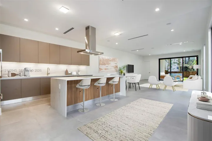 Modern kitchen featuring matte cabinetry, a waterfall-edge island with wood-finish detailing, stone-surfaced countertops, a stainless steel range hood, and large-format tile flooring