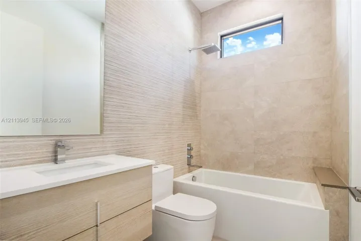 Floating vanity with light wood-finish cabinetry, rectangular under-mount sink, integrated wall-to-wall mirror, textured tile accent wall, and a bathtub with a polished chrome rain-style shower head