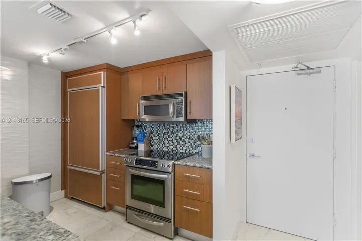 Kitchen featuring wood-veneer cabinetry, integrated stainless steel refrigerator, mosaic tile backsplash, speckled stone countertops, and stainless steel range