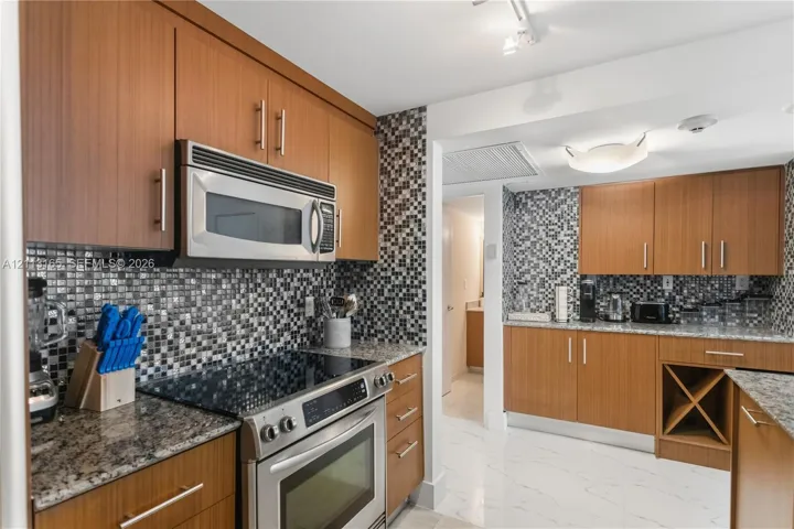 Kitchen featuring wood-finish cabinetry, stone countertops, mosaic tile backsplash, built-in wine rack, and stainless steel appliances