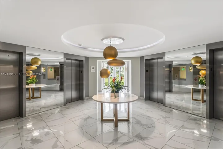 Elevator lobby featuring polished stone flooring, recessed circular ceiling detail, stainless steel elevator doors, full-height wall mirrors, and tiered metallic pendant lighting
