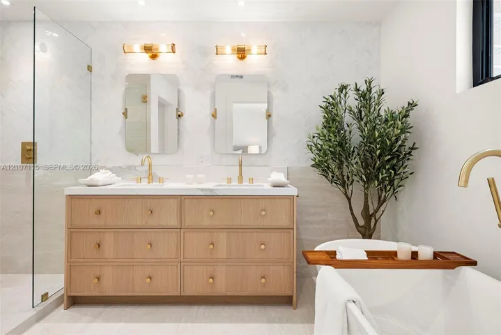 Double vanity featuring light wood cabinetry with gold-toned hardware, stone countertops, dual rectangular mirrors, and a glass-enclosed shower with herringbone wall tiling