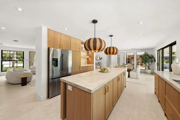Kitchen featuring wood-finish cabinetry, a large central island with light-toned countertop, stainless steel refrigerator, recessed lighting, and large-format tile flooring