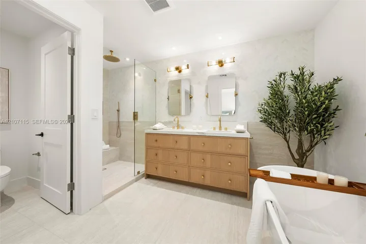 Double vanity with wood-finish cabinetry, countertop with integrated dual sinks, gold-tone wall-mounted lighting, glass-enclosed shower with tile surround, and freestanding soaking tub