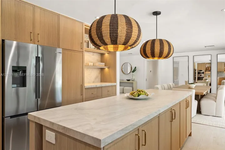 Kitchen featuring light-tone wood cabinetry, stone countertops, stainless steel refrigerator, and two hanging woven pendant lights
