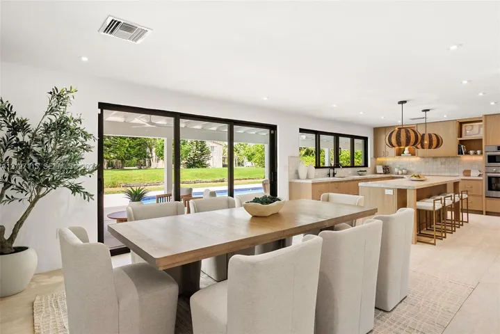 Open-concept kitchen and dining area featuring light wood-finish cabinetry, a large kitchen island with integrated seating, recessed lighting, and expansive multi-panel sliding glass doors