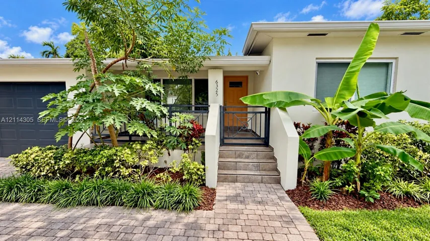 Stucco exterior featuring a paved stone walkway, covered front porch with metal railing, single-car garage, and mature landscaping