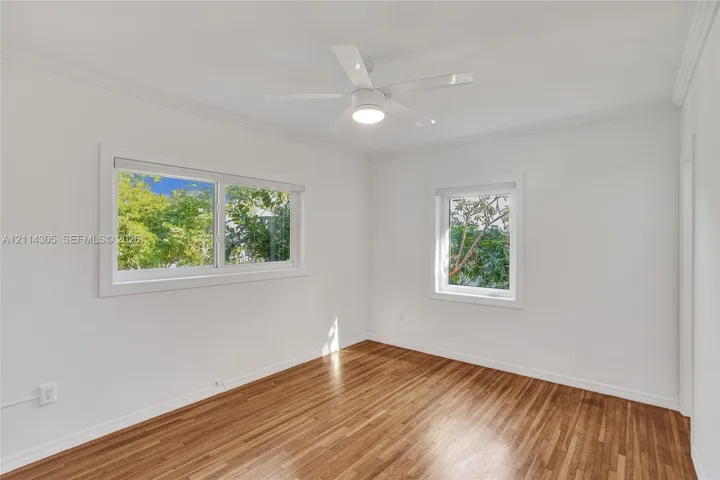 Room featuring wood-finish flooring, white walls, two windows with exterior greenery, and a white ceiling fan with integrated lighting