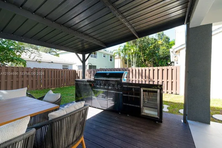 Covered patio featuring a wood-finish deck and an outdoor kitchen equipped with a stainless steel grill and a built-in beverage refrigerator, enclosed by a privacy fence